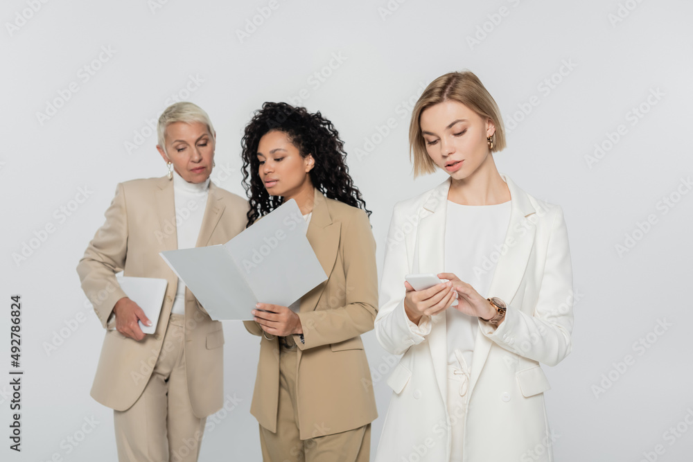 Blonde businesswoman using smartphone near interracial colleagues with paper folder isolated on grey. Blonde businesswoman using smartphone near interracial colleagues with paper folder isolated on grey.