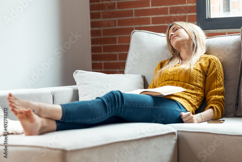 Pretty young woman falling sleep while reading a book sitting on sofa at home.
