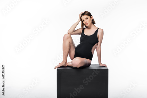 Pregnant woman in swimsuit posing on black cube isolated on white