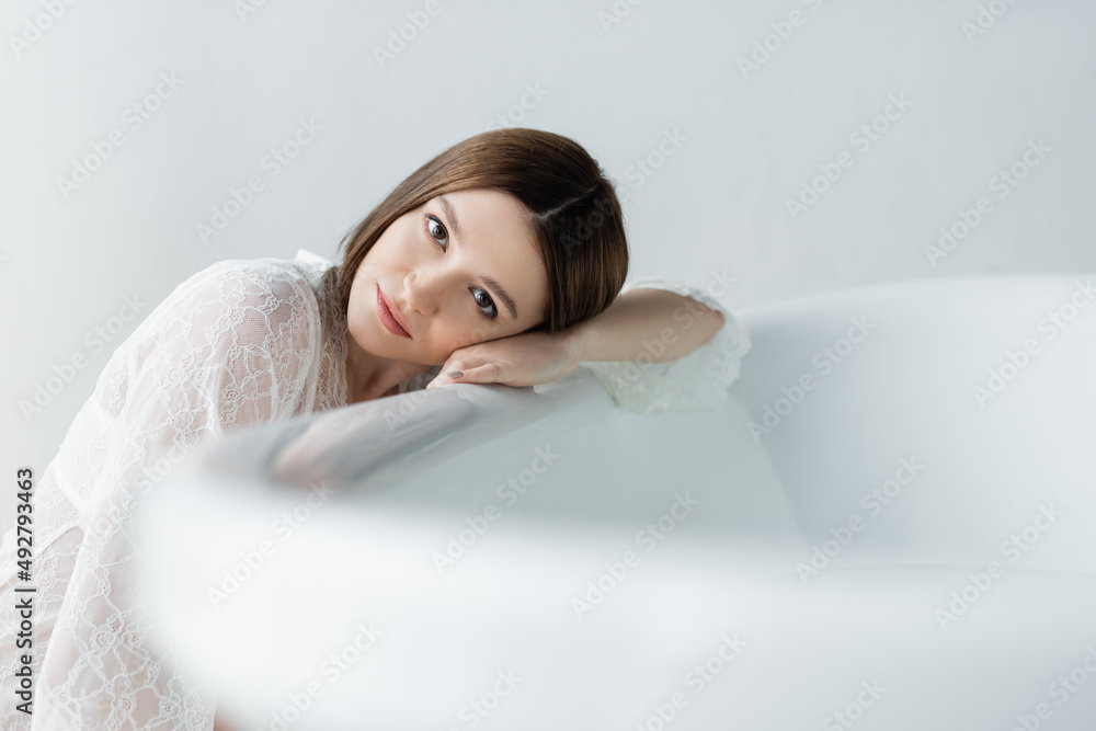 Fototapeta premium Pretty brunette woman in robe looking at camera near blurred bathtub