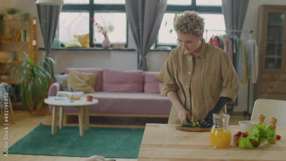 Girl with prosthetic arm cutting fresh cucumber at kitchen table and giving piece of it to dog while cooking meal at home Girl with prosthetic arm cutting fresh cucumber at kitchen table and giving piece of it to dog while cooking meal at home