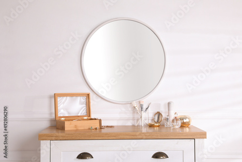 Trendy round mirror and chest of drawers with accessories near white wall indoors. © New Africa