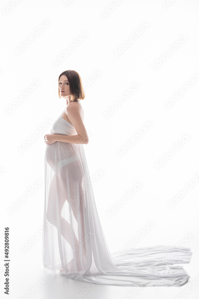 Fototapeta premium Pregnant woman in cloth and panties looking at camera on white background