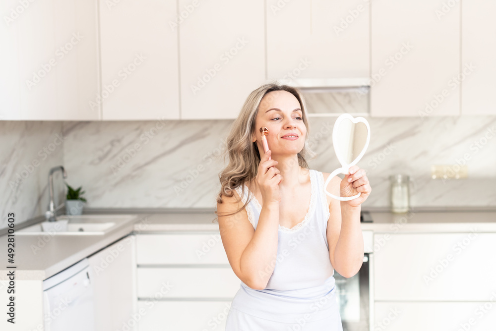 Middle age woman in pajama at kitchen with face massage tool and mirror. Beauty and anti-aging procedure for skin care. Facial care. Skin cosmetology. Morning routine, Home spa and wellness Middle age woman in pajama at kitchen with face massage tool and mirror. Beauty and anti-aging procedure for skin care. Facial care. Skin cosmetology. Morning routine, Home spa and wellness