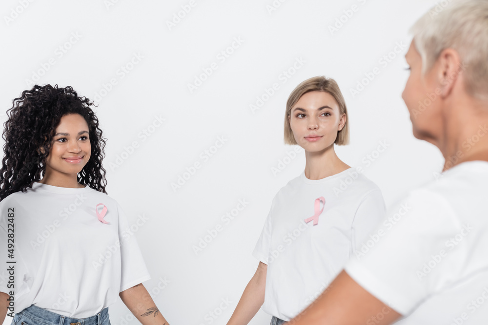 Smiling interracial women with ribbons of breast cancer awareness looking at senior friend isolated on grey. Smiling interracial women with ribbons of breast cancer awareness looking at senior friend isolated on grey.
