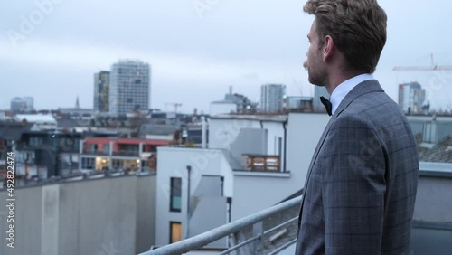 There is a man in a nice suit and a lovely mustache who is looking at his pocket watch during a cold wintersday. The grey sky looks above a small city in the backround and small snowflakes coming down photo