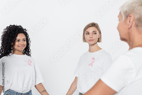 Smiling interracial women with ribbons of breast cancer awareness looking at senior friend isolated on grey.