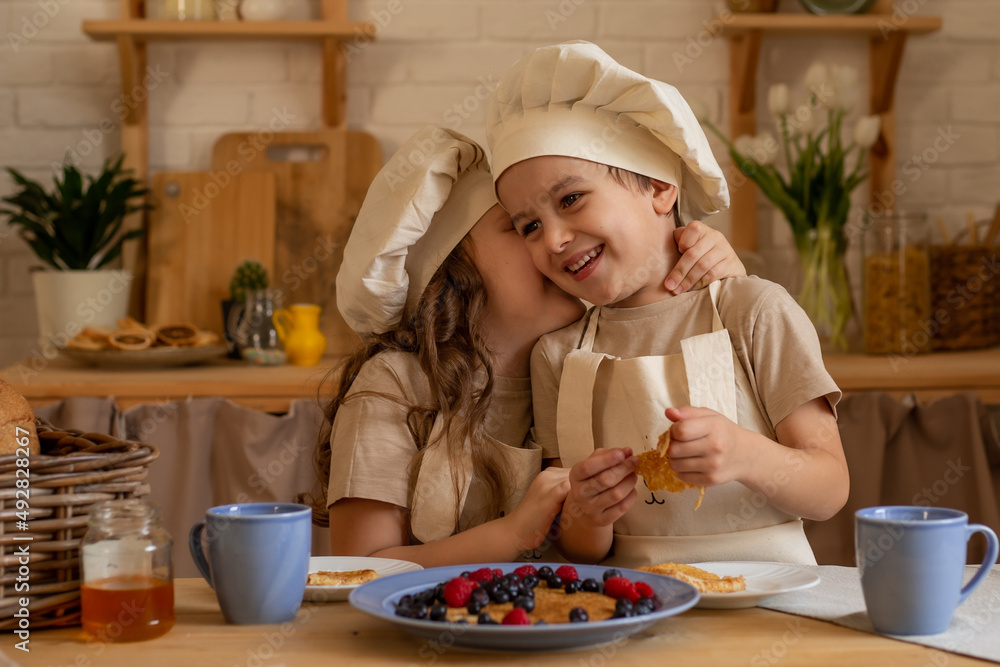 a six-year-old girl and a four-year-old boy in chef's hats and aprons are sitting at a wooden table in the kitchen a six-year-old girl and a four-year-old boy in chef's hats and aprons are sitting at a wooden table in the kitchen