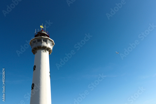 White lighthouse and white airplane in blue sky on sunny day.