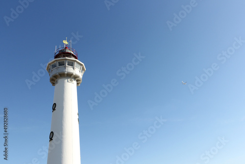 White lighthouse and white airplane in blue sky on sunny day.