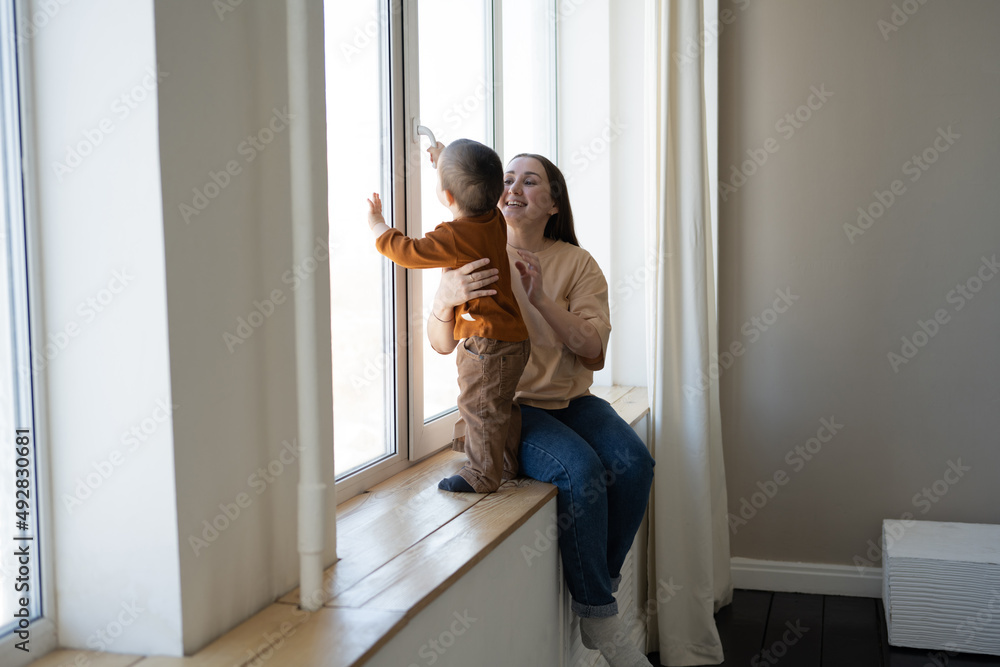 A mother and a small child stand at a large white window and look out into the street. Safety for small children in the house. - obrazy, fototapety, plakaty 