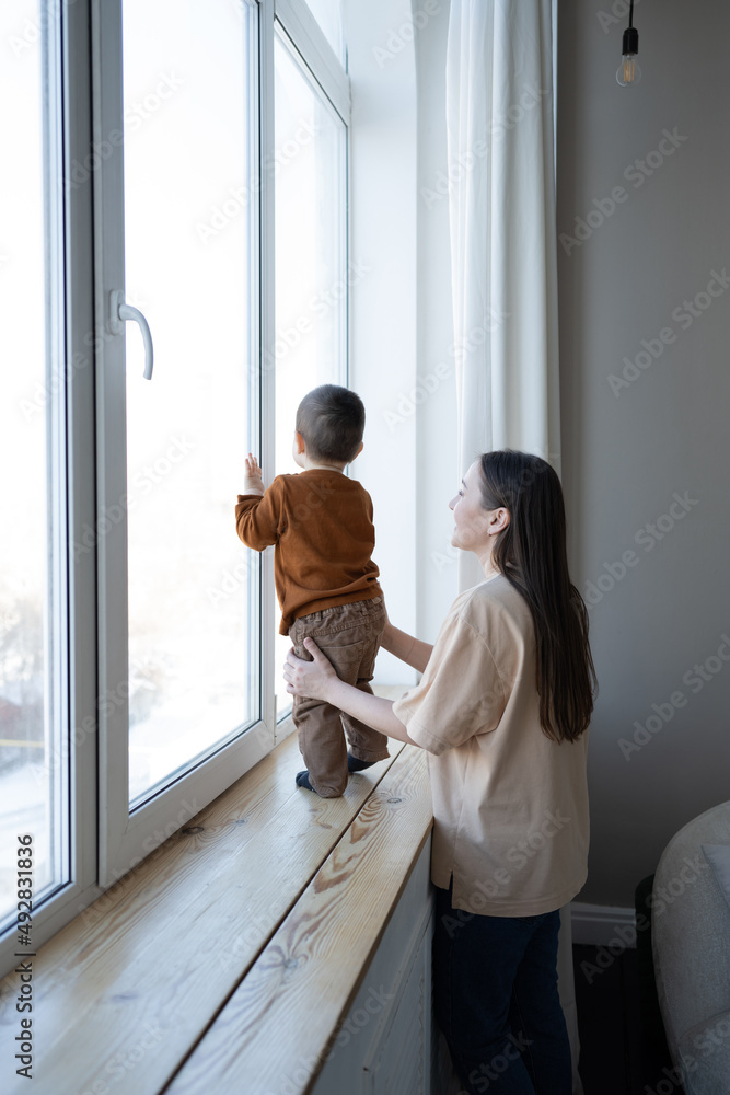 A mother and a small child stand at a large white window and look out into the street. Safety for small children in the house. A mother and a small child stand at a large white window and look out into the street. Safety for small children in the house.