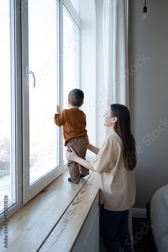A mother and a small child stand at a large white window and look out into the street. Safety for small children in the house.