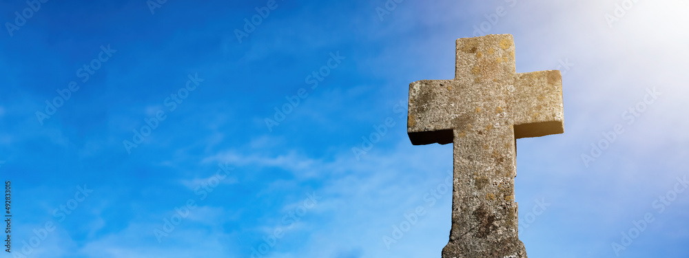 Religious Background - Blue cloudy sky withold weathered stone cross, illuminated by the sun
