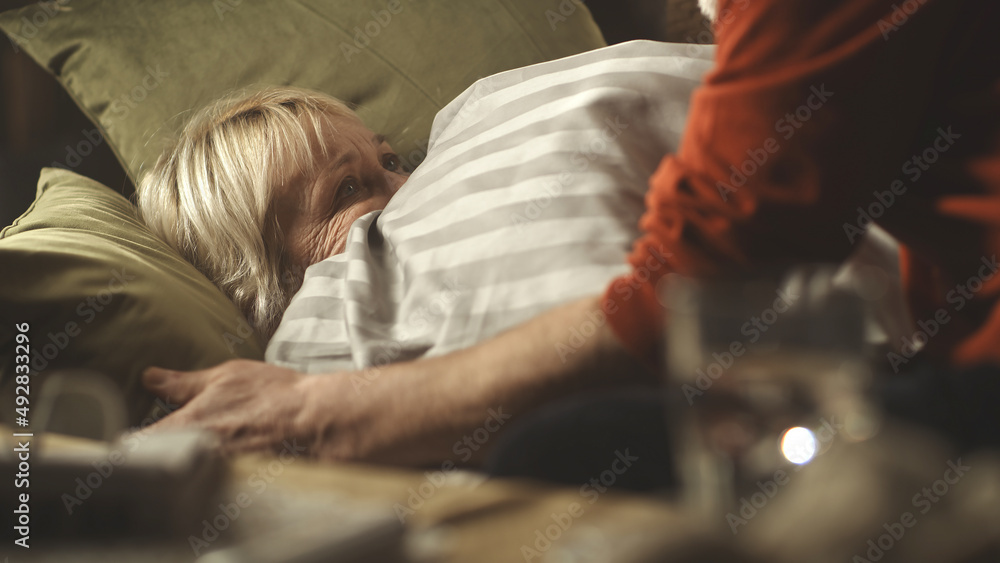 Caring senior husband in glasses giving pill and water to ill wife lying on couch in cozy living room at home Caring senior husband in glasses giving pill and water to ill wife lying on couch in cozy living room at home