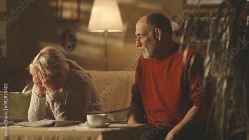 Irritated aged husband blaming upset wife while sitting on sofa at home together