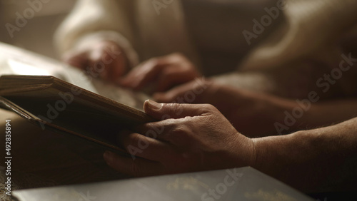 Crop senior husband and wife holding hands and flipping pages of photo album while recollecting old memories in weekend at home