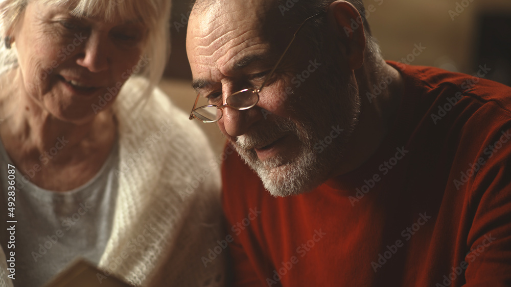 Elderly husband embracing laughing wife and speaking while examining pictures in weekend morning at home Elderly husband embracing laughing wife and speaking while examining pictures in weekend morning at home