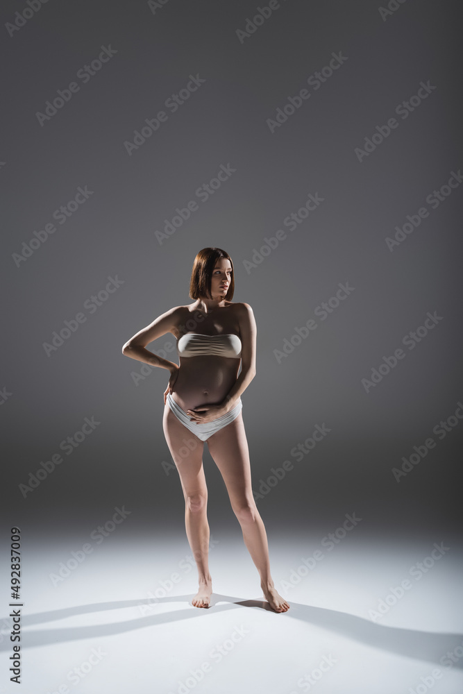Pregnant woman in underwear touching belly on grey background