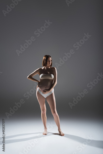 Pregnant woman in underwear touching belly on grey background