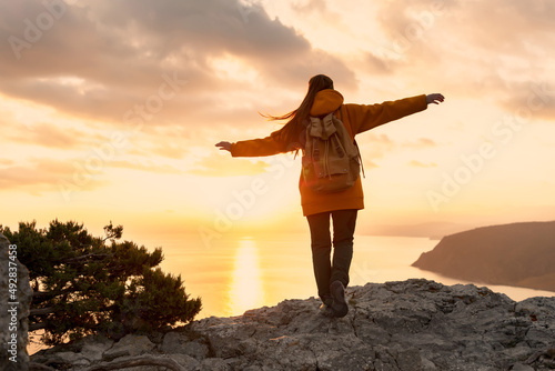 Female hiker walks with backpack on top of mountain. Girl stands with raised arms at sunset and enjoys freedom