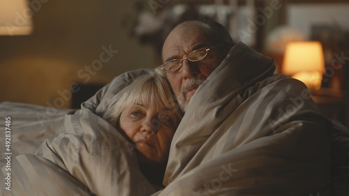 Upset elderly man discussing gas crisis and war with sad wife while wrapping in warm blanket on couch in dim cold living room at home photo