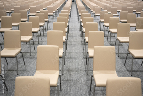 Top view of many empty cream modern chairs arranged neatly in long rows and spacing each other for social distancing  to prevent the spread of Covid-19 or Omicron  on gray carpet floor in bright hall.