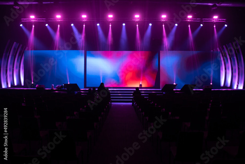 Front view of the stage event with a big LED screens and purple and pink lights are shining down on the stage floor while testing the light system with many empty chairs arranged for audience in hall. photo