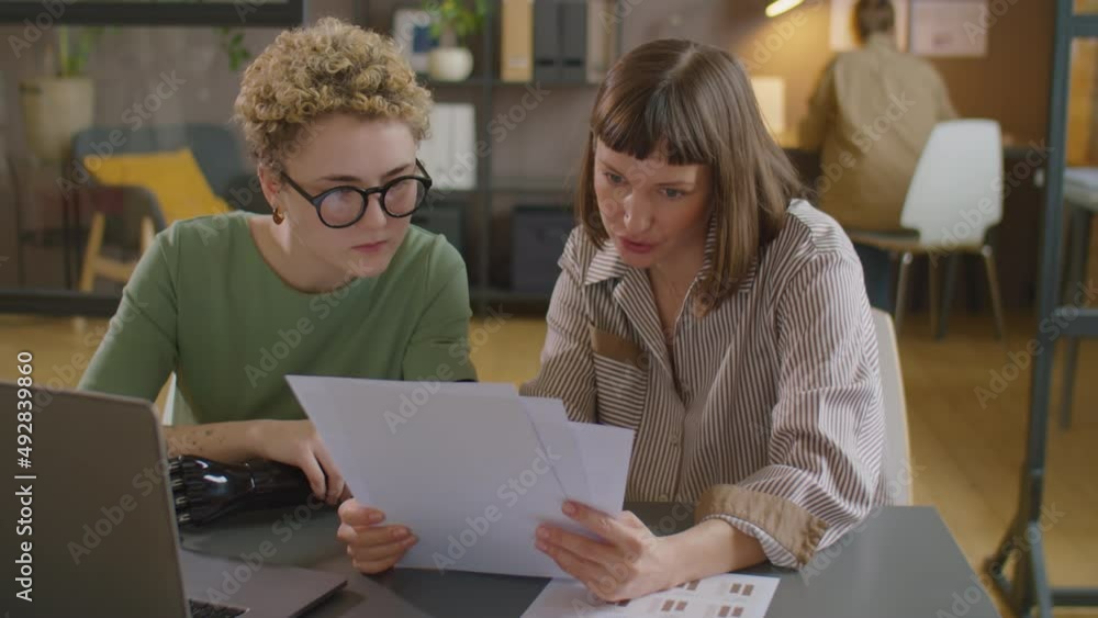 Young businesswoman with prosthetic arm and her female colleague sitting at desk, discussing papers and using laptop while working together on business project in office Young businesswoman with prosthetic arm and her female colleague sitting at desk, discussing papers and using laptop while working together on business project in office