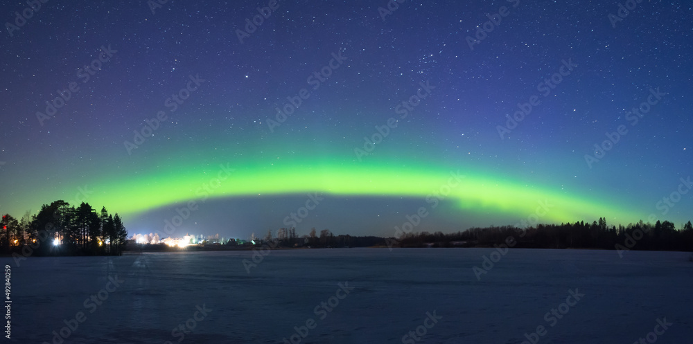 Polar northen lights aurora borealis at night in the starry sky above the lake with the island and the silhouette of the trees by the forest. Glowing arc over the horizon, panorama.