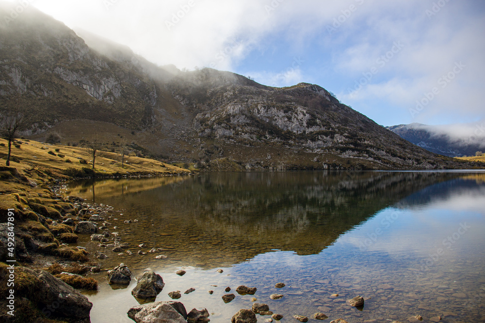 Fototapeta premium Paisaje montañoso con neblina, adornado en la orilla de un lago pedregoso. Cielo azul y reflejos del lago .Lagos de Covadonga 