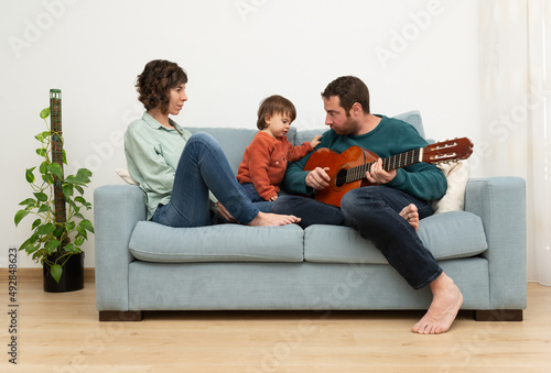 Parents and toddler sitting on a sofá having time together. Man is playing a spanish guitar while his little girl and wife are looking him and enjoying the music. 