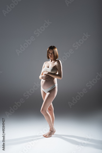 Barefoot pregnant woman in underwear looking at camera on grey background