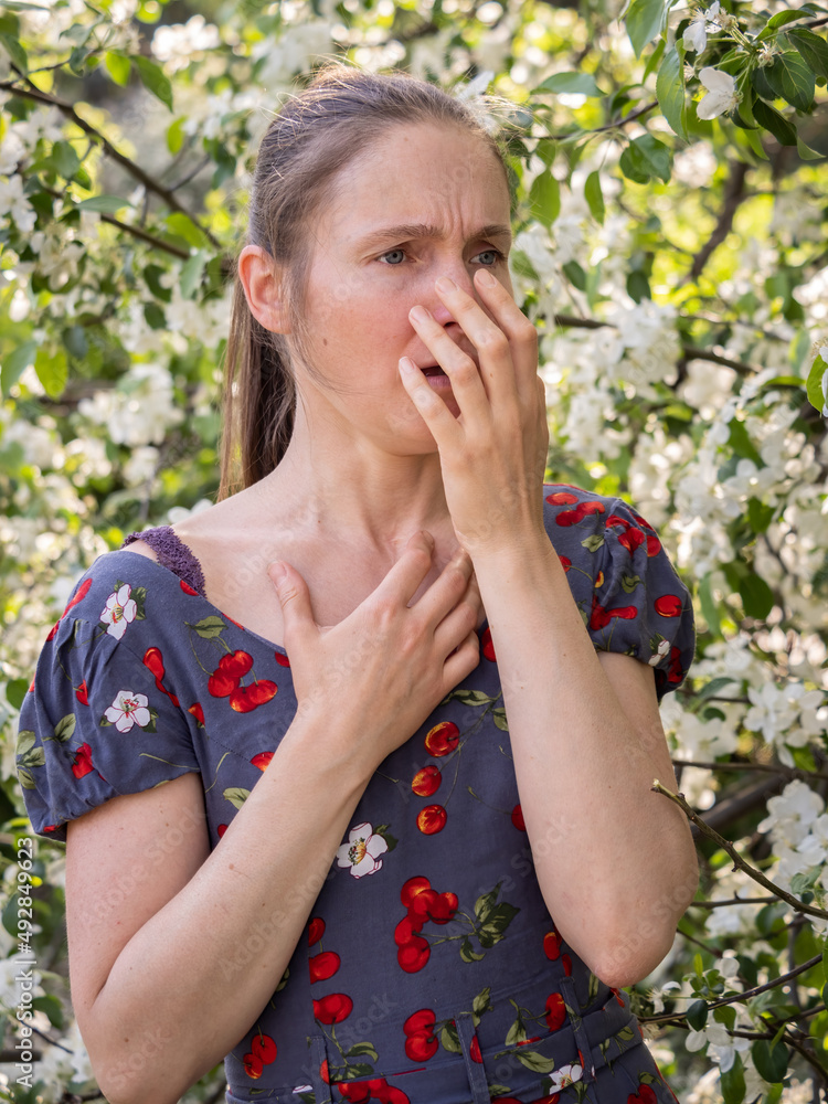 Young woman sneezes in the park against the background of a flowering tree. Allergy to pollen concept. Young woman sneezes in the park against the background of a flowering tree. Allergy to pollen concept.