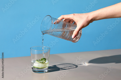 Minimal close up of female hand pouring water into glass against blue background, hydration and detox concept, copy space