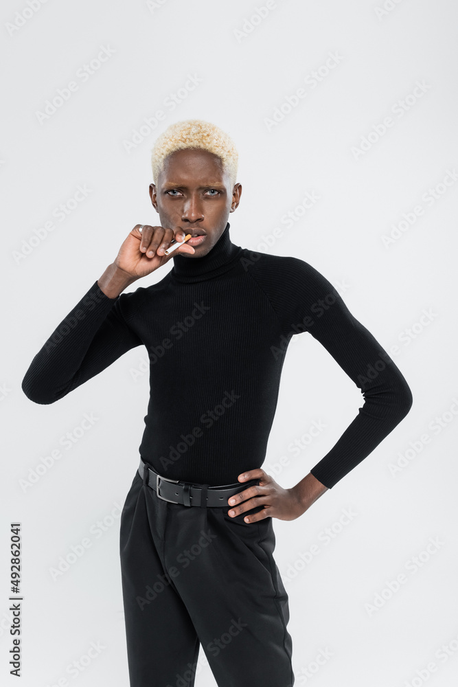 young and blonde african american man holding cigarette and standing with hand on hip isolated on grey young and blonde african american man holding cigarette and standing with hand on hip isolated on grey