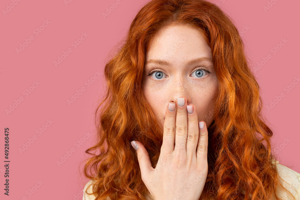Close up photo of a redhead girl with blue eyes looking at the camera with surprised facial expression covering her mouth with a palm. Close up photo of a redhead girl with blue eyes looking at the camera with surprised facial expression covering her mouth with a palm.