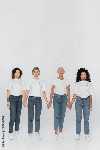 Multiethnic women with ribbons of breast cancer awareness holding hands on grey background.