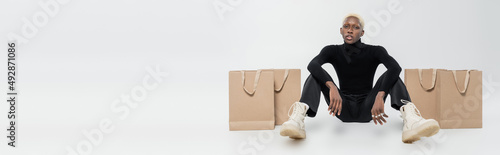 full length of blonde african american man sitting near paper bags on grey, banner