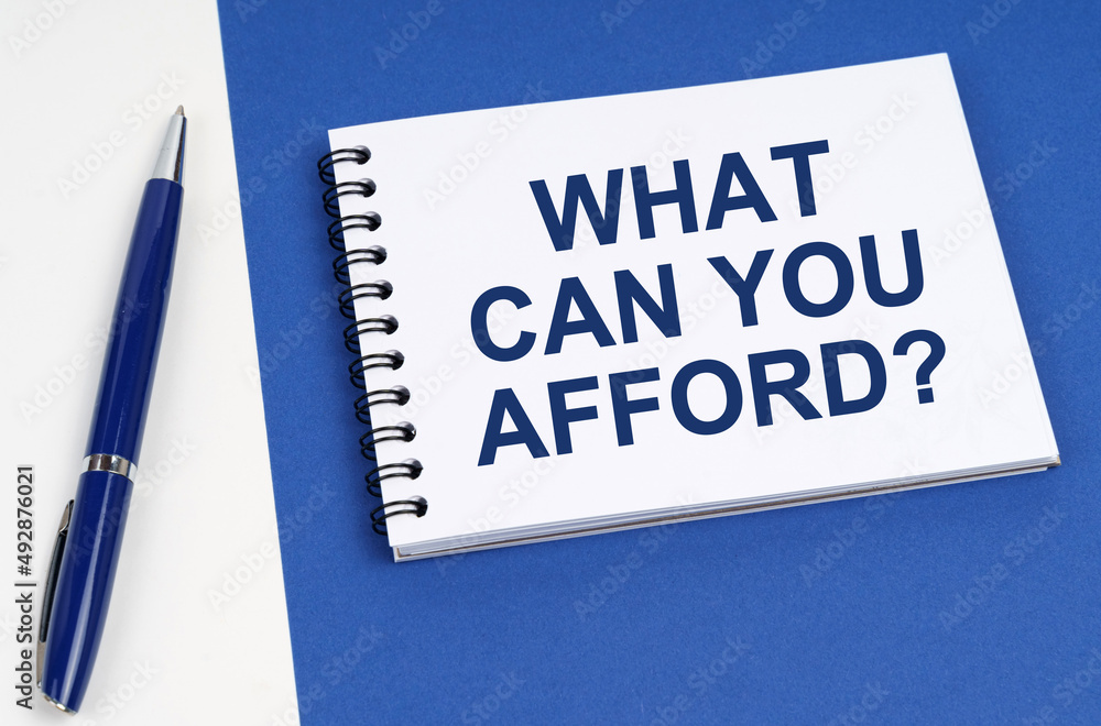 On a blue-white surface lies a pen and a notebook with the inscription - What Can You Afford On a blue-white surface lies a pen and a notebook with the inscription - What Can You Afford