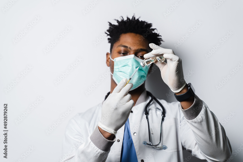 African doctor in a medical mask prepares to give an injection of a coronavirus covid-19 vaccine. Black man in medical mask is making vaccination on white background.