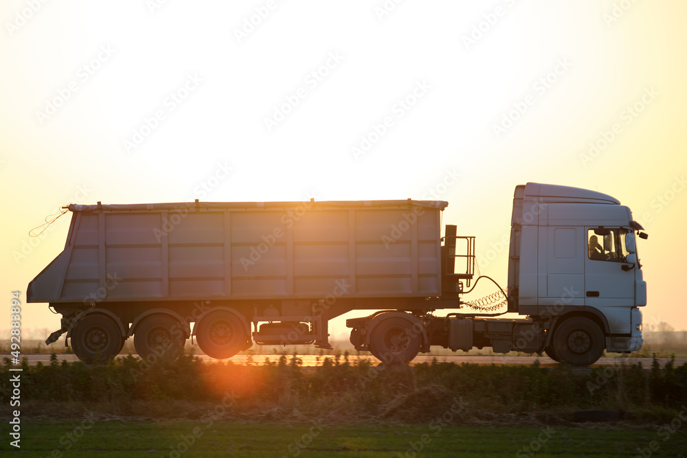 Semi-truck with tipping cargo trailer transporting sand from quarry driving on highway hauling goods in evening. Delivery transportation and logistics concept