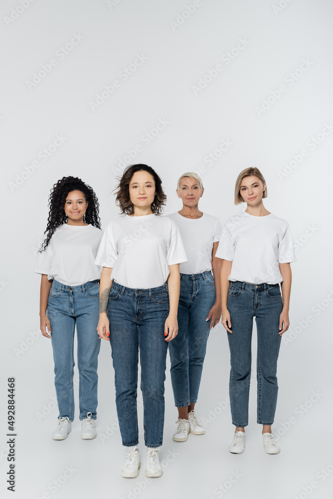 Full length of smiling multiethnic women in white t-shirts standing on grey background. Full length of smiling multiethnic women in white t-shirts standing on grey background.