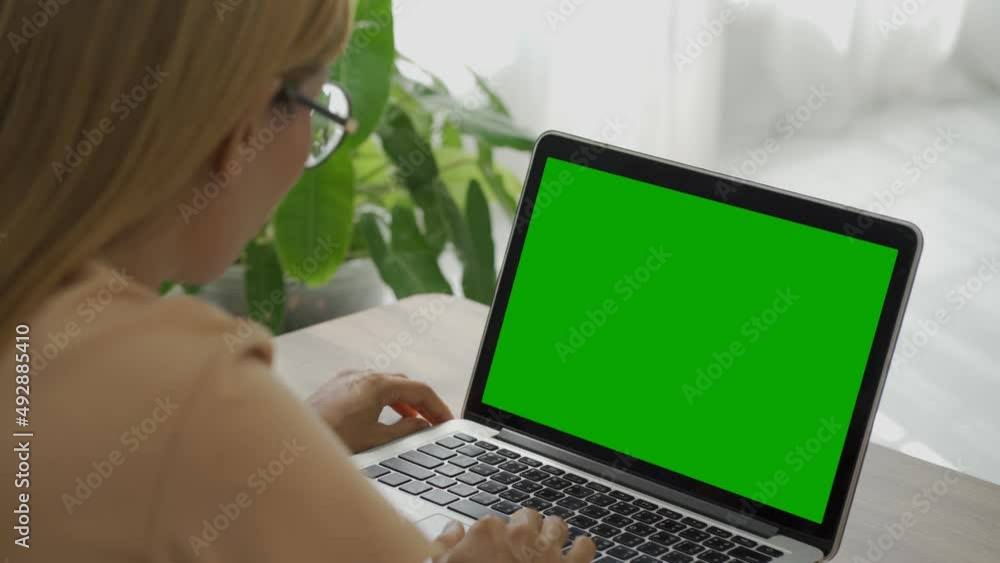 Over the shoulder shot of a business woman working in office interior on pc on desk, looking at green screen. Office person using laptop computer with laptop green screen.