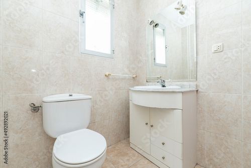 bathroom with white porcelain fixtures  cream tile walls  wooden cabinet with marble top and frameless mirror with sconces