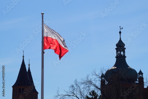 The white and red Polish flag in the wind against the blue sky and historic buildings in Płock, Poland.