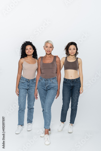 Full length of multiethnic women standing near elderly friend on grey background.