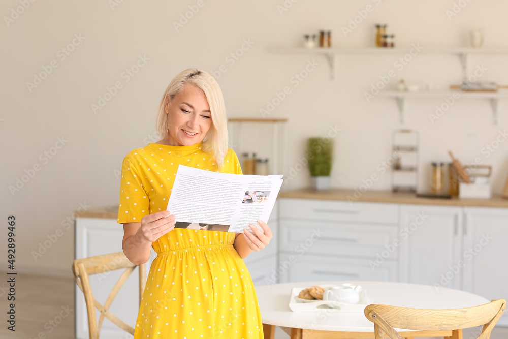 Beautiful mature woman reading newspaper in kitchen