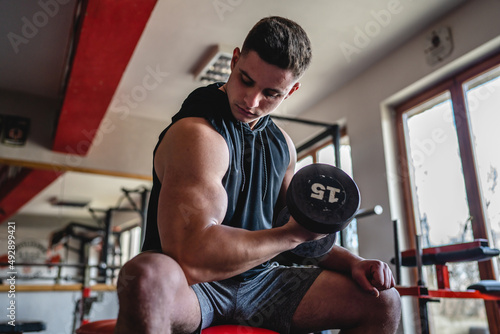 One man young adult caucasian male bodybuilder training arms bicep flexing muscles with dumbbell while sitting in the gym wearing shirt real people copy space front view