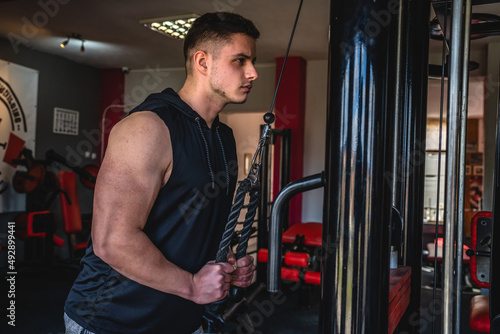 One man side view caucasian male bodybuilder at gym wearing black shirt workout using rope and cable weights for arms exercise triceps training copy space waist up dark photo real people amateur
